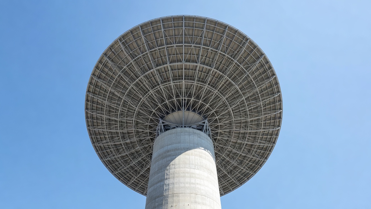 Over-the-shoulder view of a radio telescope dish array on an arid high-altitude plateau, concrete support structures visible, pointing toward a pale midday sky, shot on 85mm lens, f/8, bright high-key daylight, harsh overhead sun casting sharp shadows.