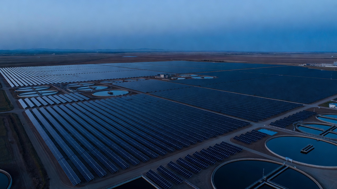 Aerial establishing view of the 27 MW-DC solar array spread across graded plots at Fresno's Northeast Surface Water Treatment Plant, cool blue-hour sky, long exposure, wide lens.