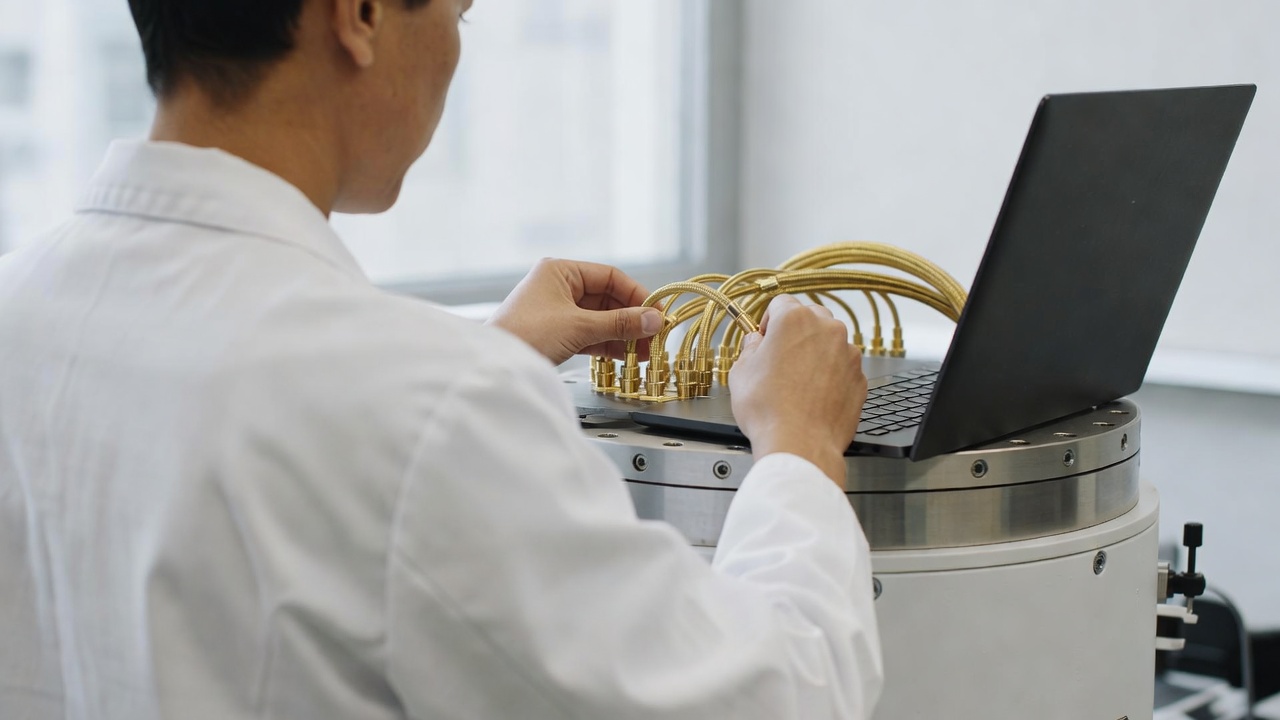 Researcher in a white lab coat connects gold coaxial cables to the top plate of a cryostat, with a laptop beside the cables.
