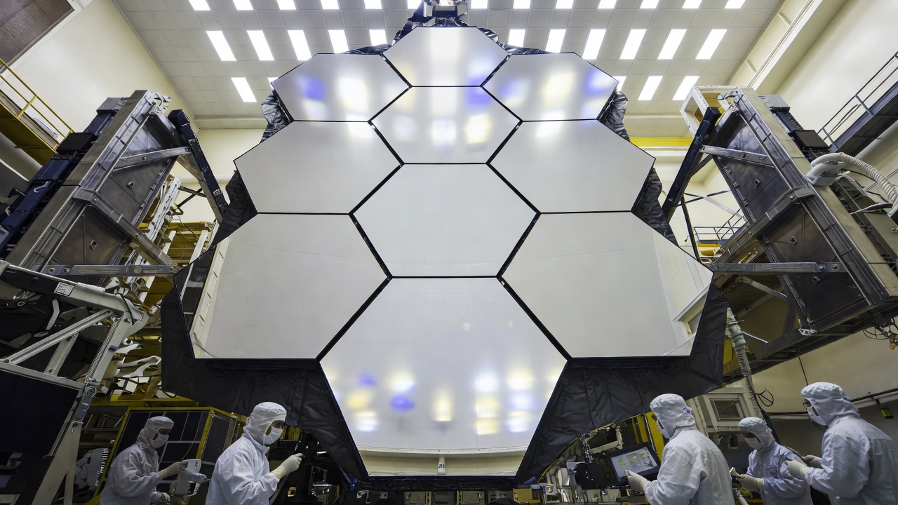 interior of a large-format space telescope mirror assembly inside a cleanroom facility, technicians in white suits visible at the periphery, no text, no labels, no watermarks