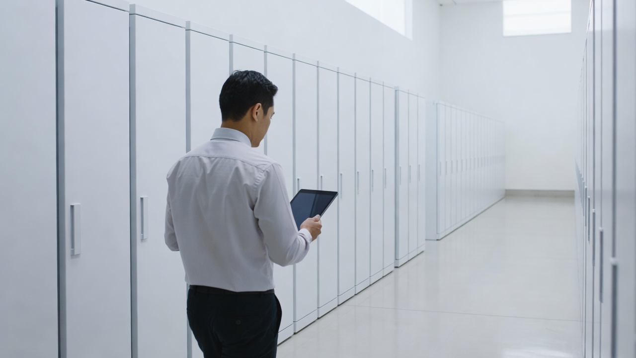 Engineer in a light shirt seen from behind, holding a tablet while walking along a long row of tall white equipment cabinets in a bright hall.
