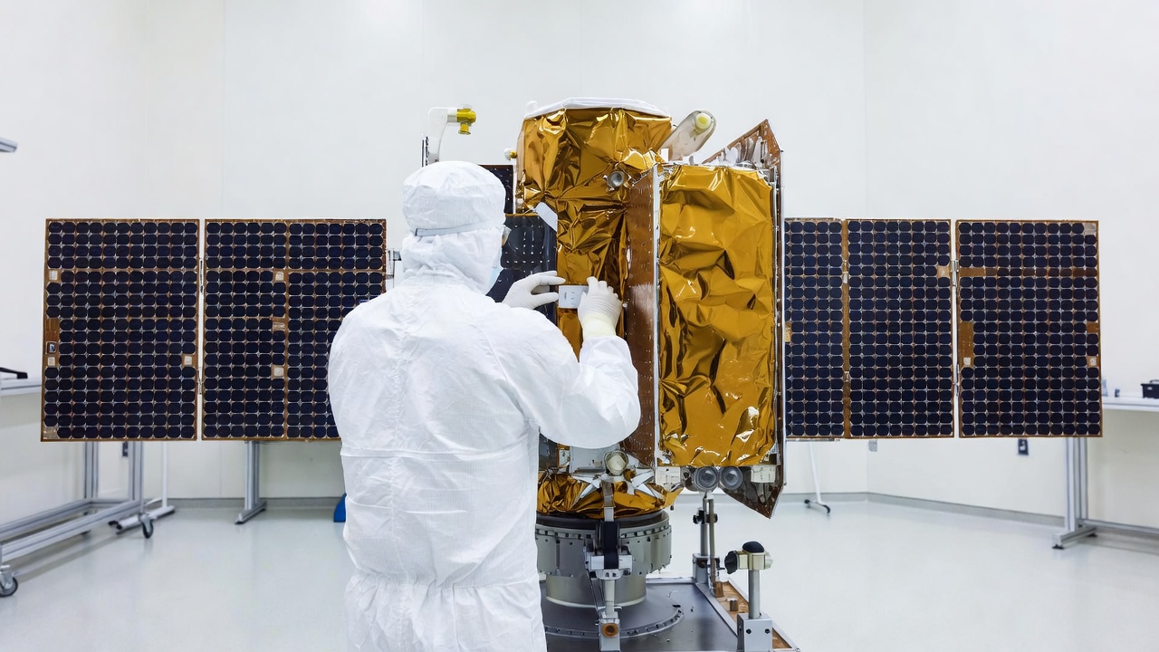 Technician in a white cleanroom suit, facing away, works on a gold-foil-wrapped satellite with extended solar panels inside a bright cleanroom.