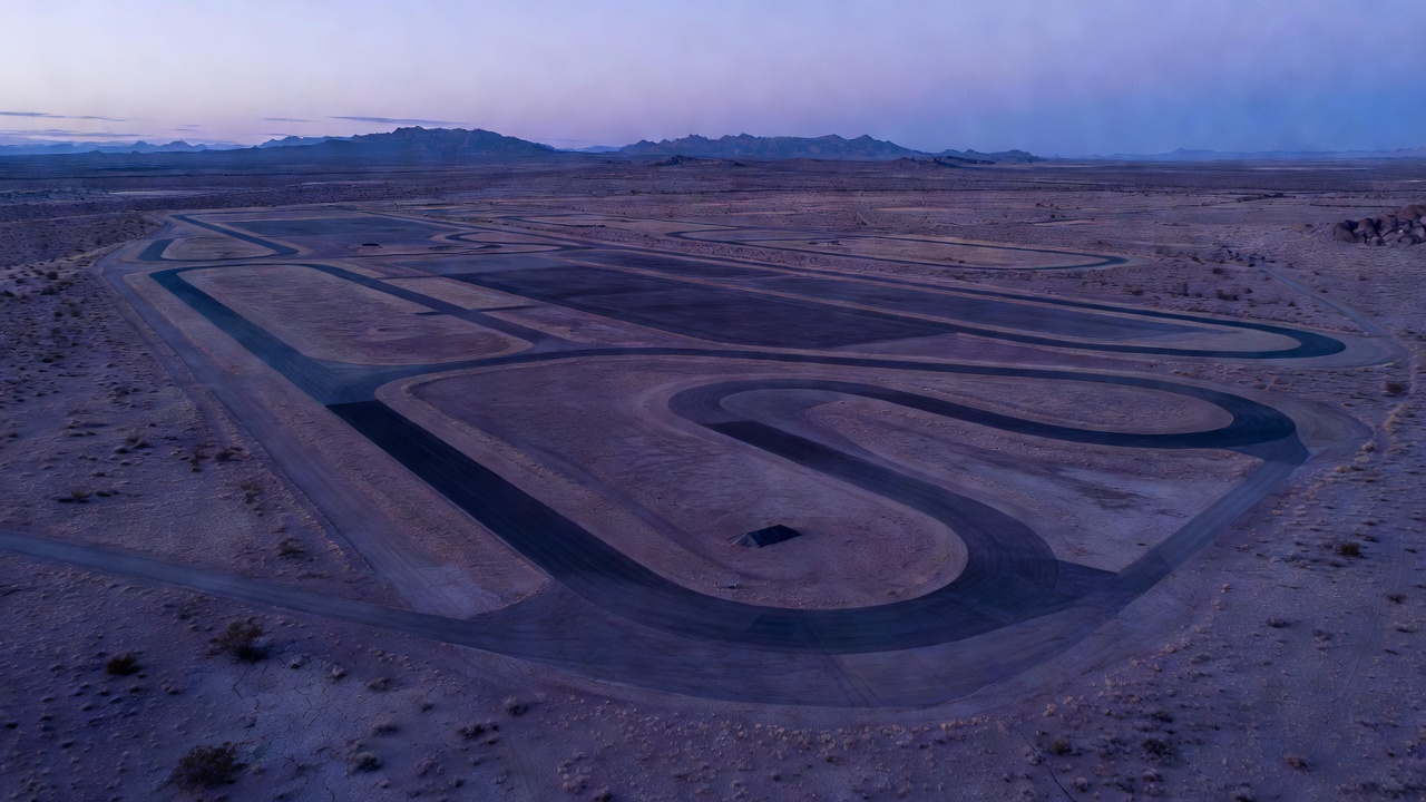 Aerial wide-angle view of a vast 5,500-acre desert test facility in Arizona at blue hour, empty asphalt proving grounds stretching toward distant mountains, cool twilight tones, drone-perspective wide lens.