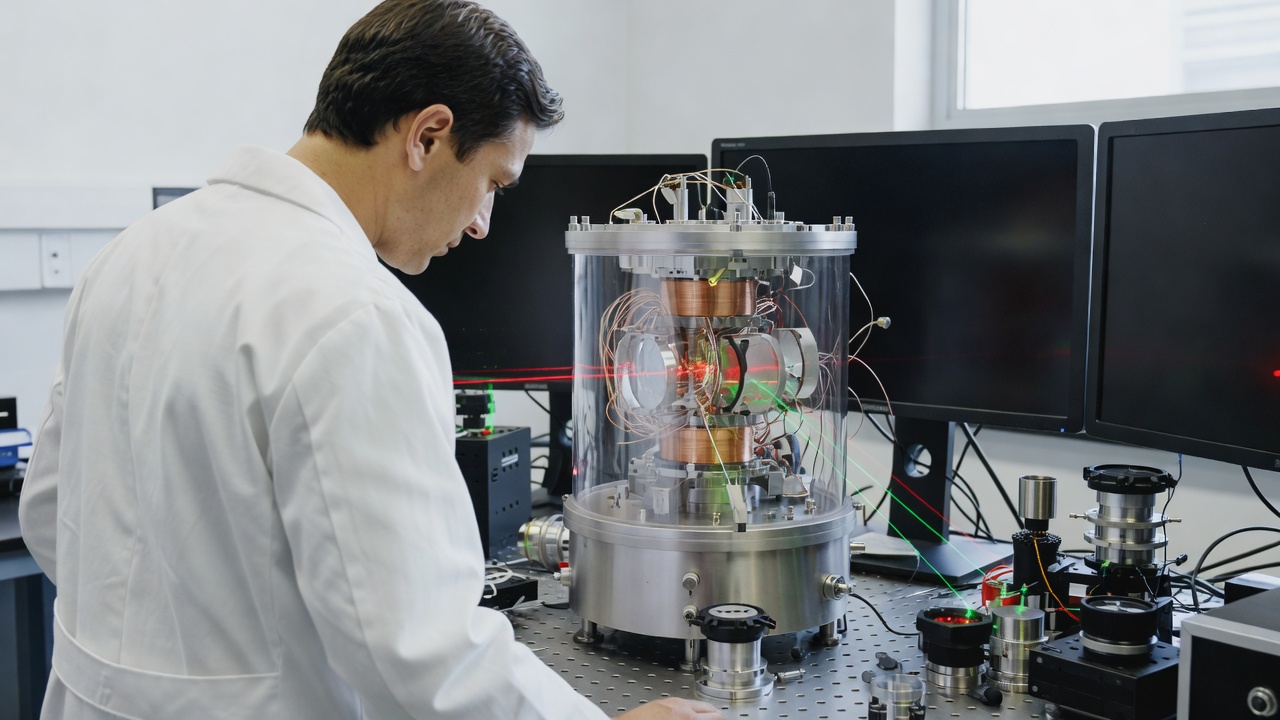 Scientist in a white lab coat studies a glass vacuum chamber with copper coils and red and green laser beams on an optical table.