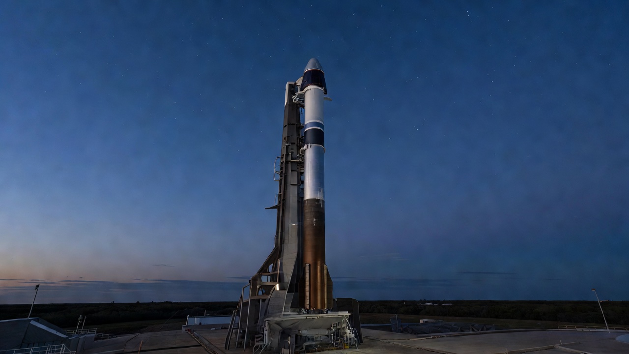 Tall white and blue rocket with a capsule-shaped nose stands on a launch pad beneath a deep blue twilight sky with faint stars.