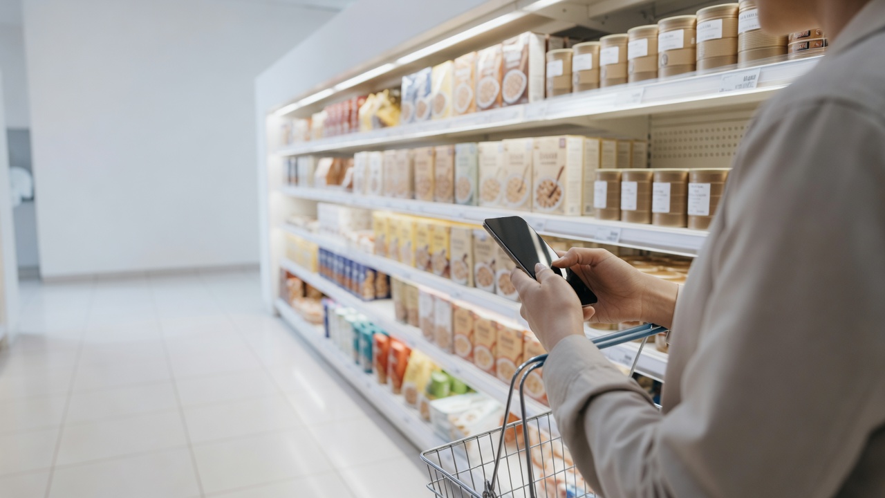 Shopper's hands hold a smartphone in a bright supermarket aisle lined with boxed cereal and tinned goods, a wire basket at their side.