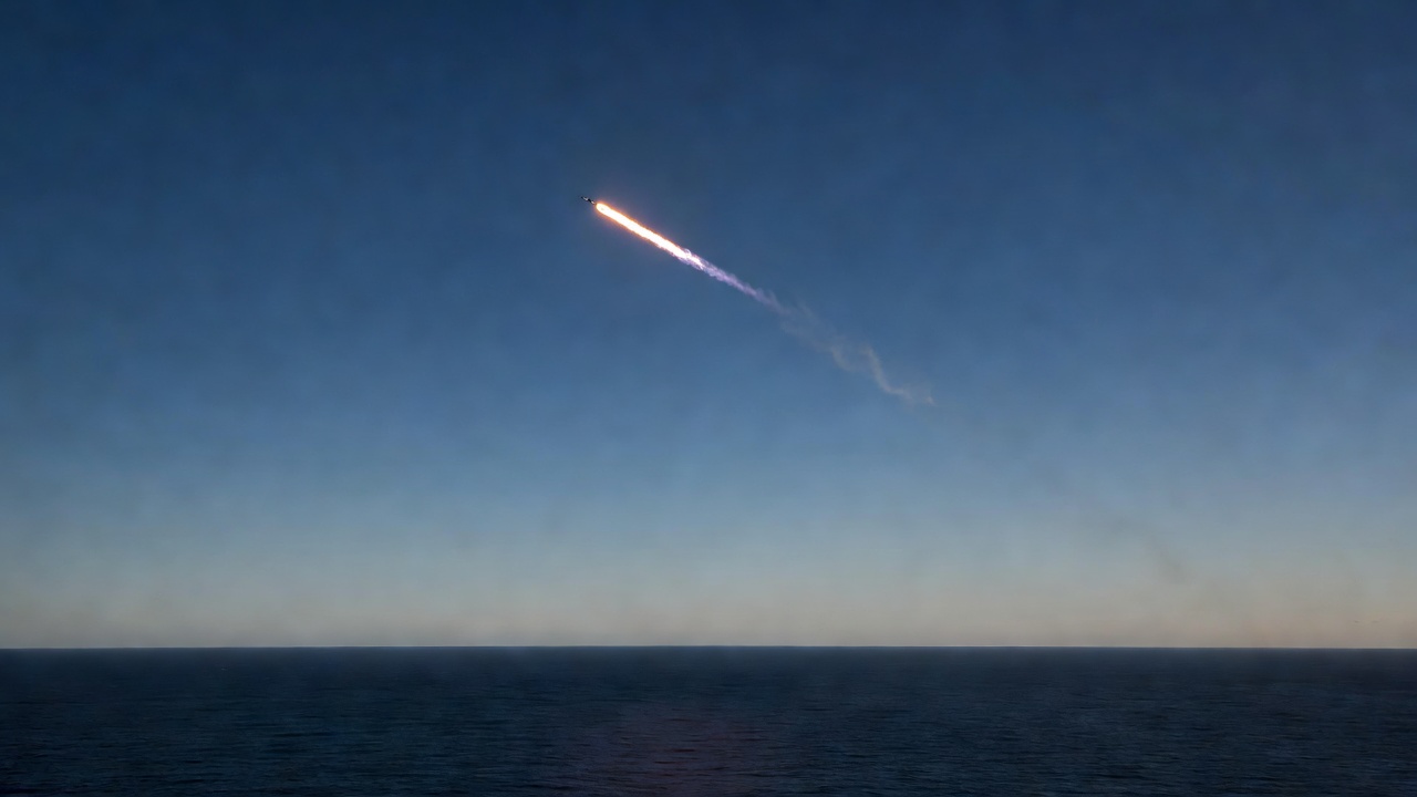 A rocket's glowing exhaust streak and thin trailing plume arc across a deep blue twilight sky above a calm, dark ocean horizon.