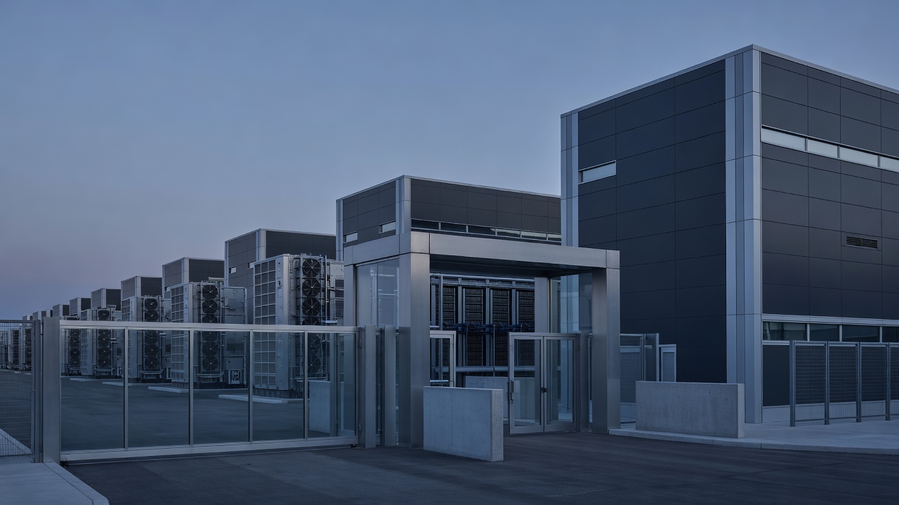 Wide shot of a modern data center exterior at blue hour, rows of cooling units and server building facades visible, enterprise security checkpoint gate in foreground, cool twilight sky casting blue-grey light across the complex, long focal length compression.