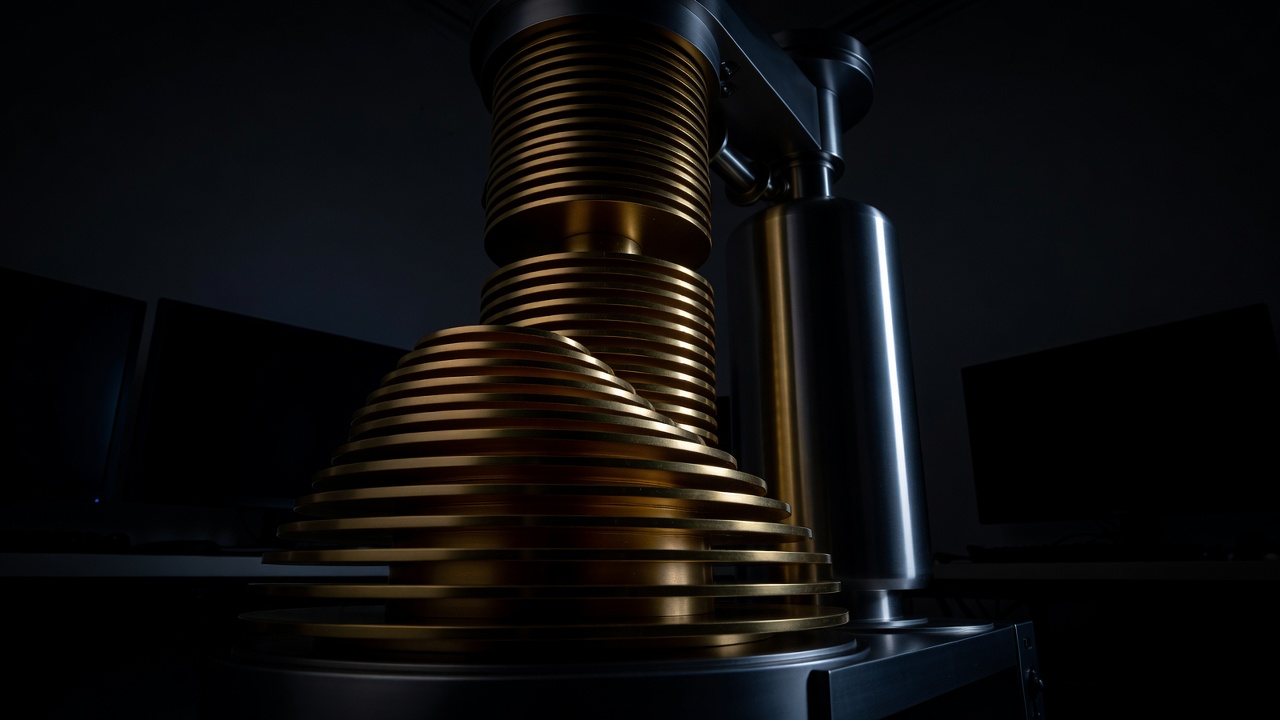 A gold tiered quantum cooling apparatus and stainless steel cylinder in a dark lab with monitors silhouetted behind.