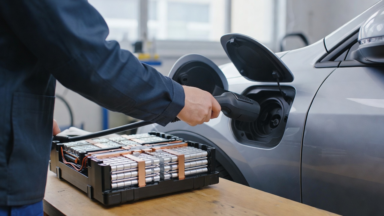 Technician's hand guiding a charging connector into a car's open charge port in a garage, with a battery pack module on the workbench.